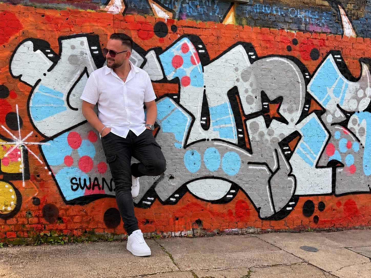 G Prospex wearing sunglasses and a white shirt in front of a colourful graffiti wall